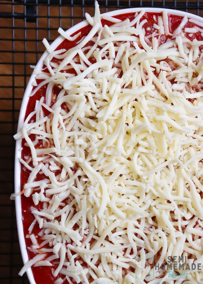 adding shredded cheese to baked ravioli with sauce in a white casserole dish