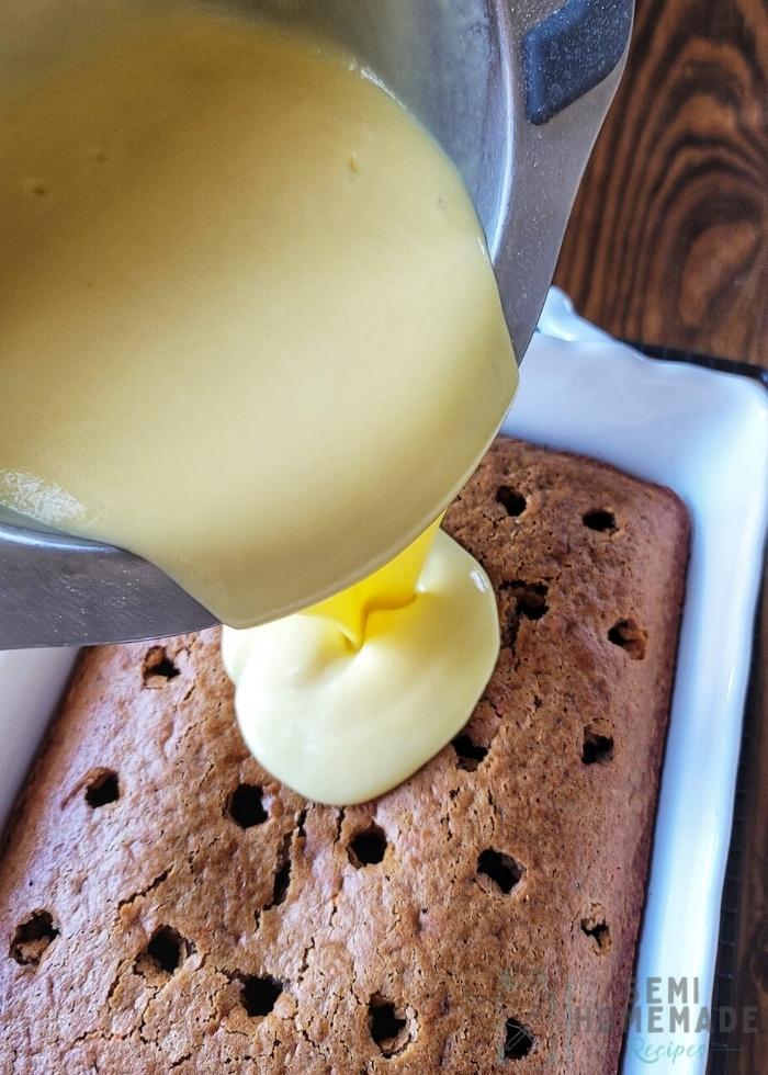 pouring vanilla pudding onto carrot cake with holes in a white baking dish
